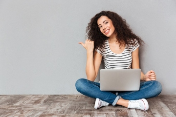 Nice woman with beautiful smile sitting in lotus pose on the floor with silver computer on legs gesturing thumb aside, submitting something copy space