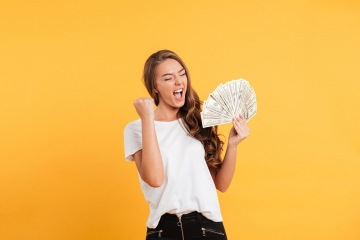 Portrait of a pleased joyful girl holding bunch of money banknotes and celebrating isolated over yellow background