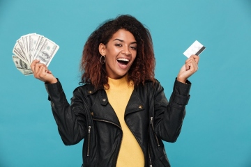 Happy african woman in leather jacket holding money and credit card while looking at the camera over blue background