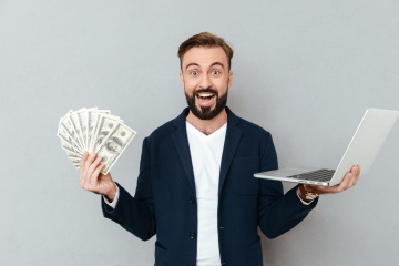 Happy surprised bearded man in business clothes holding money and laptop computer while looking at the camera over gray background