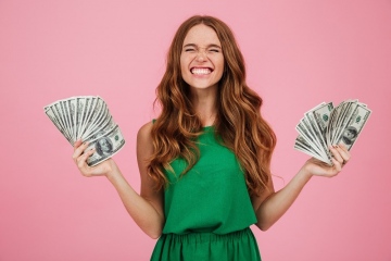 Portrait of a satisfied happy woman winner with long hair holding bunch of money banknotes in each hand isolated over pink background